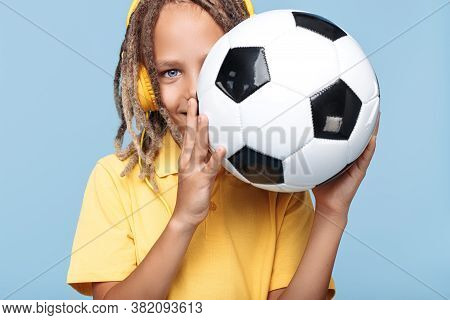 Cheerful Little Boy With African Dreads Cover Face With Soccer Ball Over Blue Background.