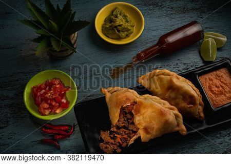 Traditional baked Latin American empanadas savoury pastries with meat beef stuffing  with tomato and avocado sauces on blue wooden background. Top view. 