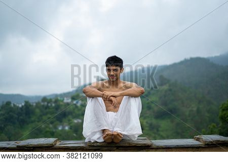 Young Shredded Man Sitting Wearing A Dhoti And Glasses With Blurred Mountains In The Background.