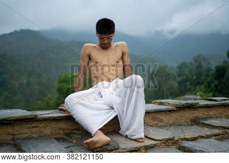 Young Shredded Man Sitting Wearing A Dhoti And Glasses With Blurred Mountains In The Background.