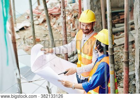 Positive Builders In Yellow Hardhats Checking Building Blueprint And Discussing Plan Or Workrs
