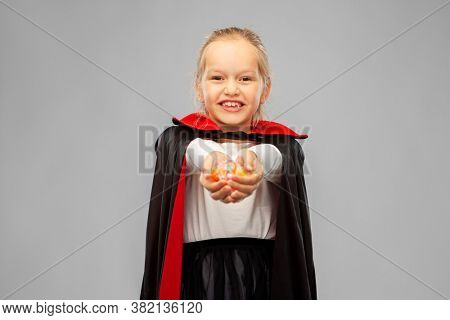halloween, holiday and childhood concept - happy smiling girl in black dracula cape or costume with candies over grey background