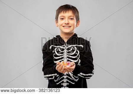 halloween, holiday and childhood concept - smiling boy in black costume of skeleton with candies over grey background