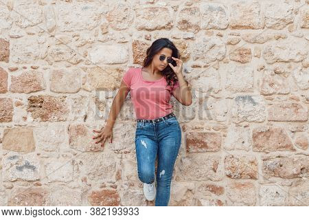 Young Women Wearing T-shirt And Jeans Stays Near A Wall And Touch Sunglasses
