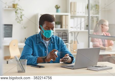 Portrait Of Young African-american Man Wearing Mask Sanitizing Hands While Sitting At Desk In Cubicl