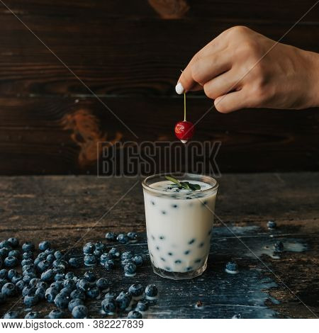 A Womans Hand Holds A Cherry And Soaks It In Milk. Blueberries Are Scattered