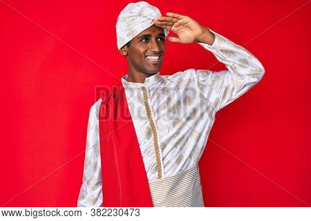 Handsome indian man wearing tradition sherwani clothes very happy and smiling looking far away with hand over head. searching concept. 