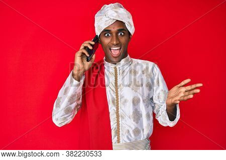 Indian man wearing tradition sherwani clothes speaking on the phone celebrating achievement with happy smile and winner expression with raised hand 