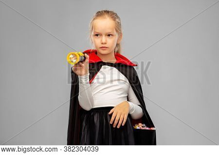 halloween, holiday and trick-or-treating concept - girl in black dracula cape or costume with candies and flashlight over grey background