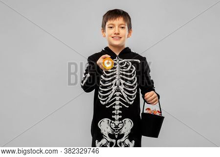 halloween, holiday and trick-or-treating concept - smiling boy in black costume of skeleton with candies and flashlight over grey background