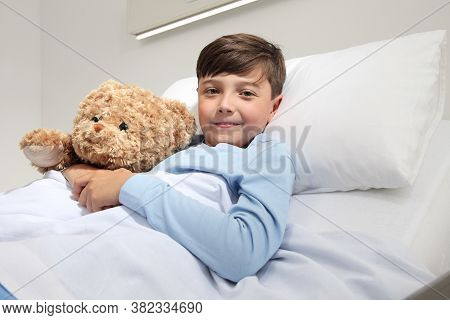 Portrait Of Cute Happy Child Alone In Hospital Room Lying In Bed With Teddy Bear Looking At Camera A