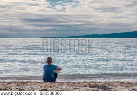 A Young Boy Sitting On The Sand At The Beach Overlooking The Sea And An Island. This Is In St Helier