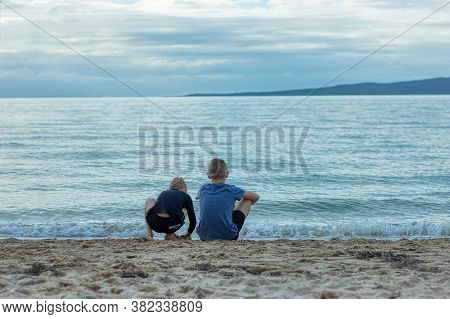 Two Young Brothers Sitting On A Beach In St Heliers, Auckland, New Zealand. The Older Brother Is Loo
