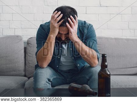 Despair And Alcoholism. Sad Man Alcoholic Holds His Head With Hands, Empty Bottles On Table In Inter