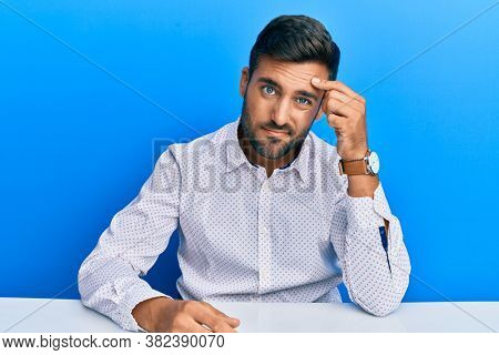 Handsome hispanic man wearing business clothes sitting on the table pointing unhappy to pimple on forehead, ugly infection of blackhead. acne and skin problem 