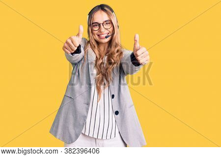 Young beautiful blonde woman wearing call center agent headset approving doing positive gesture with hand, thumbs up smiling and happy for success. winner gesture. 