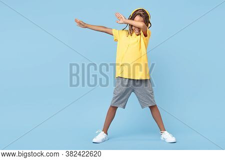 Cheerful Little Boy With African With Headphones Dabbing With One Leg On Soccer Ball Over Blue Backg