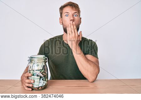 Young caucasian man holding jar with savings sitting on the table covering mouth with hand, shocked and afraid for mistake. surprised expression 