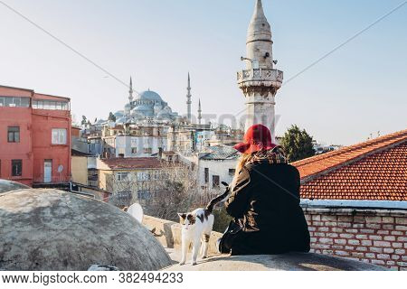 Blond Woman Stroking A Cat On The Roof With A View To The Mosque, Istanbul, Turkey. Girl In A Hat Si