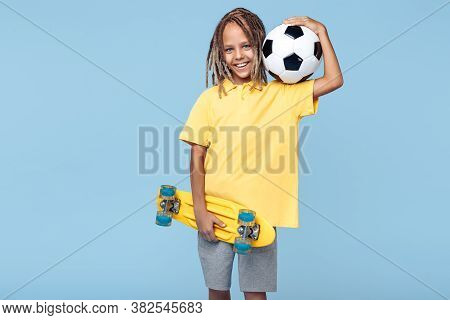 Cheerful Little Boy With African Dreads Holding Soccer Ball And Skateboard Over Blue Background.