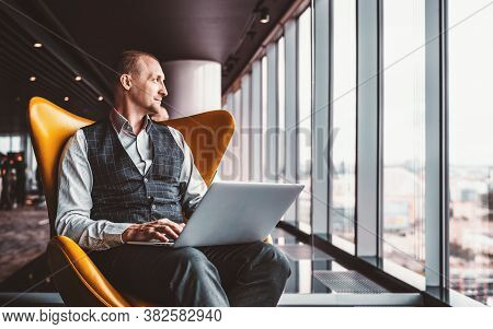 A Handsome Cheerful Caucasian Man Entrepreneur Is Sitting With A Laptop On His Knees On An Orange Ar