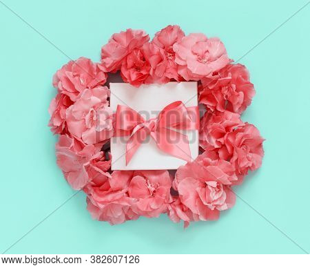 White Gift Box With A Bow Between Pink Flowers Top View On Light Green Background