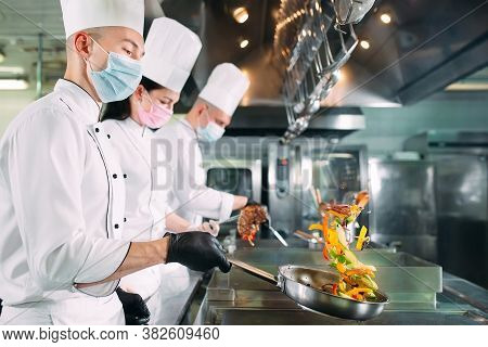 Chefs In Protective Masks And Gloves Prepare Food In The Kitchen Of A Restaurant Or Hotel.