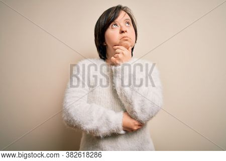 Young down syndrome woman standing over isolated background with hand on chin thinking about question, pensive expression. Smiling with thoughtful face. Doubt concept.