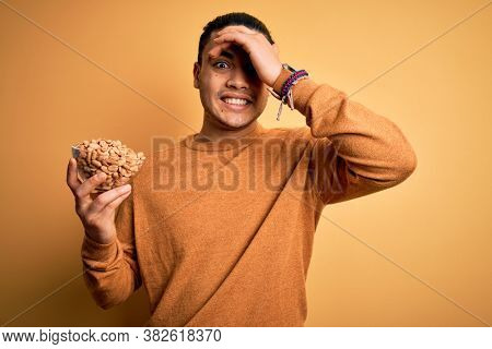 Young brazilian man holding bowl with healthy salty peanuts over isolated yellow background stressed with hand on head, shocked with shame and surprise face, angry and frustrated. Fear and upset