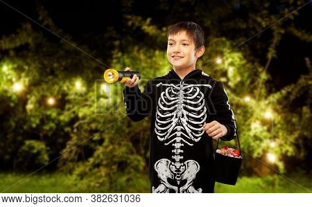 halloween, holiday and trick-or-treating concept - smiling boy in black costume of skeleton with candies and flashlight over garland lights at night park on background