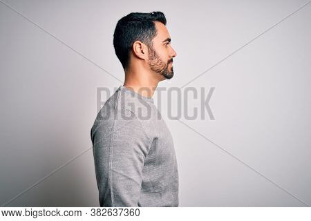 Young handsome man with beard wearing casual sweater standing over white background looking to side, relax profile pose with natural face with confident smile.