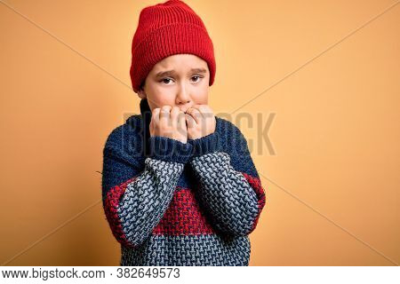 Young little boy kid wearing wool cap and winter sweater over yellow isolated background looking stressed and nervous with hands on mouth biting nails. Anxiety problem.