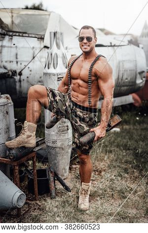 Muscular American Soldier With Stationary Airplane Behind. Brutal Man With Large Biceps And Naked To