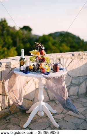 A Round Table Covered With A White Tablecloth With Champagne Glasses And Fruit By A Stone Fence Over