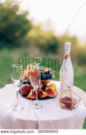 A Table With A White Tablecloth And Two Champagne Glasses And A Bookcase With Fruits In Nature.