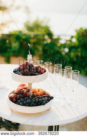 Two-tier Grocery Shelf With Grapes And Fruits On A White Table With Empty Champagne Glasses.