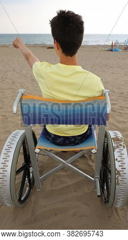 Disabled Boy In A Wheelchair Points To The Sea From The Beach
