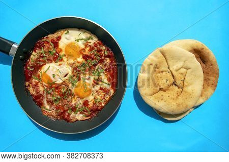 Shakshuka - Poached Eggs In Tomato Sauce, Onion, Pepper And In Iron Pan With Pita Bread On Blue Brig