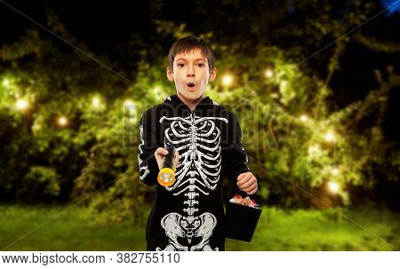 halloween, holiday and trick-or-treating concept - excited boy in black costume of skeleton with candies and flashlight over garland lights at night park on background