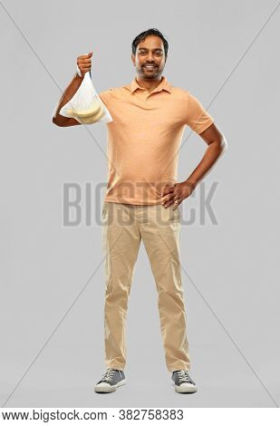 food, sustainability and eco living concept - smiling young man in striped t-shirt putting bananas into reusable string bag over grey background