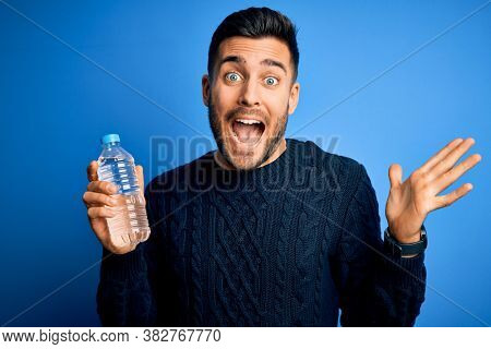 Young handsome man drinking bottle of water to refeshment over blue background very happy and excited, winner expression celebrating victory screaming with big smile and raised hands