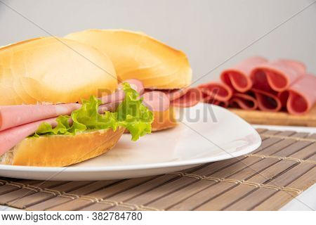 Mortadella Sandwich And Lettuce On A White Plate On A Mat And Slices Of Mortadella Next To It On A C