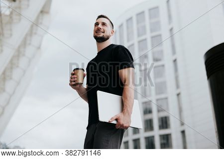 Young Handsome Smiling Man Walking Down The Street, Carrying His Laptop While Drinking Coffee On The