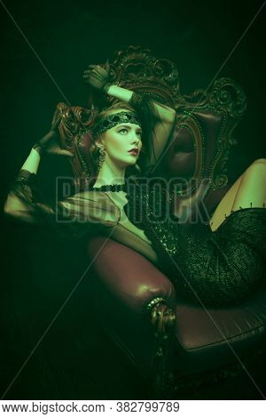 Beautiful woman in an elegant evening dress in an armchair in a rich classic interior. Clothing, makeup and hairstyle in the style of the 1920s.