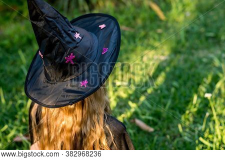 Causcasian Blond Girl With Long Hair In Halloween Witch Costume Sitting On A Green Grass
