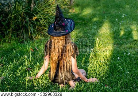 Causcasian Blond Girl With Long Hair In Halloween Witch Costume Sitting On A Green Grass.