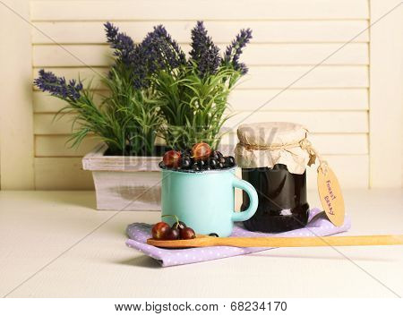 Ripe blackcurrants in mug and glass jar with tasty jam on light wooden background.
