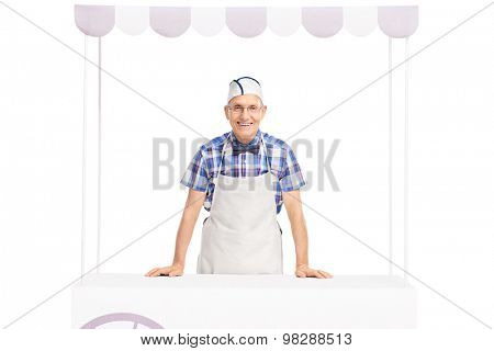 Senior ice cream seller with a white cap and apron standing behind an ice cream stand and looking at the camera isolated on white background
