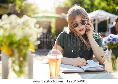 Portrait of a young business woman or student writing her plans in notepad talking on a smartphone wearing glasses during a luch break in park on a sunny summer day. Copy space.