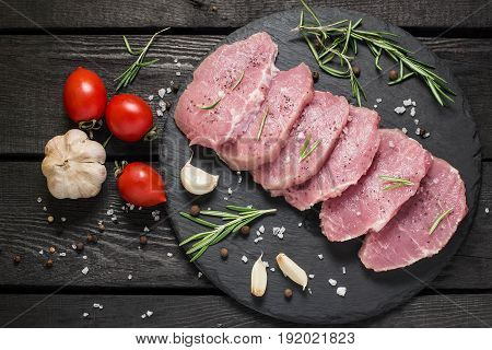 Fresh uncooked boneless pork chops on a slate plate vegetables herbs and spices on a dark wooden background. Meat prepared for cooking